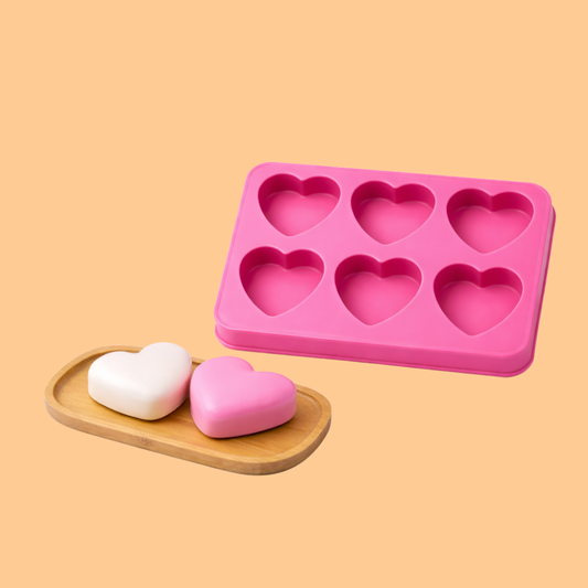Pink silicone heart shaped soap mould tray displayed beside finished heart shaped handmade soap bars on a wooden tray, flexible mold used for DIY soap making, melt and pour soap crafting, artisan soap production, and decorative cosmetic bath products.