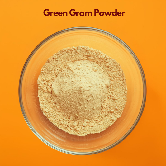 Overhead view of Green Gram Powder in a clear glass bowl on a vibrant orange background, displaying finely milled light yellow beige herbal powder with soft texture. Natural herbal powder used in face packs, gentle exfoliation treatments and cosmetic skincare blends.