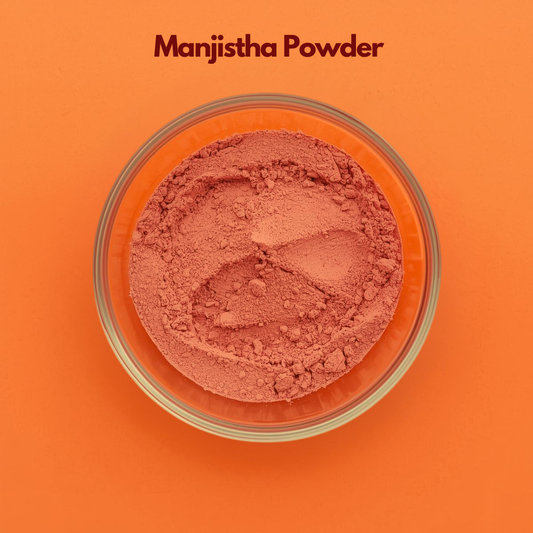Top view of Manjistha Powder in a transparent bowl, showing soft reddish brown finely ground herbal powder against a vibrant orange surface. Traditional Ayurvedic herb powder used in skin detox face packs, natural skincare blends and herbal cosmetic applications.